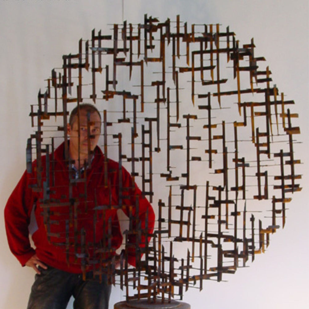 A man wearing a red sweater and dark jeans stands beside a large circular metal wall sculpture made of intersecting rods, photographed against a plain light background.